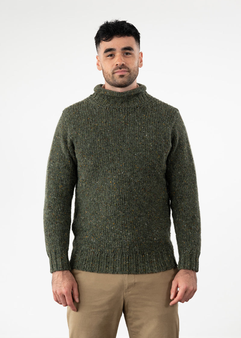 A man wearing a dark olive green knit turtleneck jumper and beige chino trousers, standing against a plain white studio background.