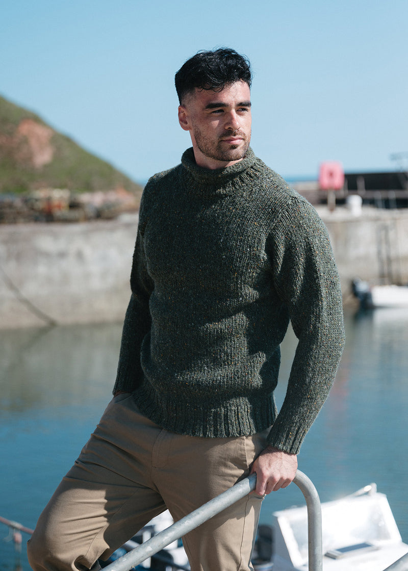 A man wearing a dark olive green knit jumper and khaki trousers, leaning on a boat railing with a harbour and water in the background.