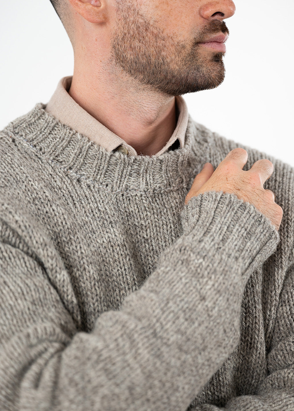 Close-up of a man wearing a chunky knit grey-brown jumper over a beige collared shirt, with his hand resting on his chest against a plain light background.