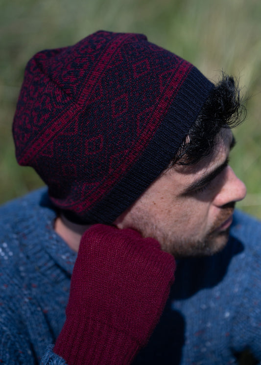 A close-up side profile of a man wearing a dark navy knit beanie with a burgundy geometric pattern and a matching burgundy knit glove, set against a blurred outdoor background.