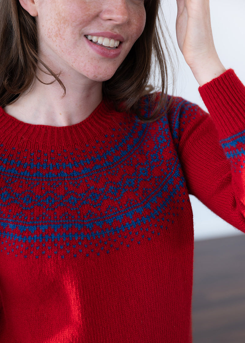 A close-up of a woman wearing a red knit sweater with a blue Fair Isle pattern across the chest, shoulders and cuff.