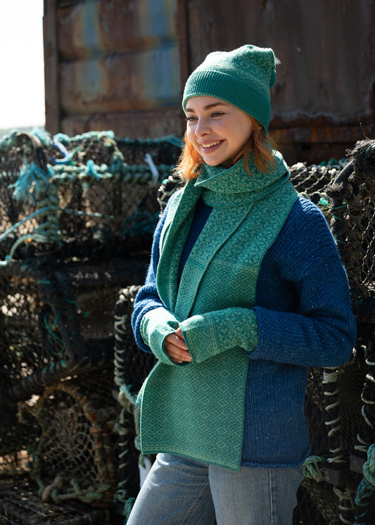 A woman wearing a sea green knit patterned scarf, matching pom pom hat, and wrist warmers, paired with a blue jumper and light blue jeans, standing outdoors in front of stacked fishing creels.