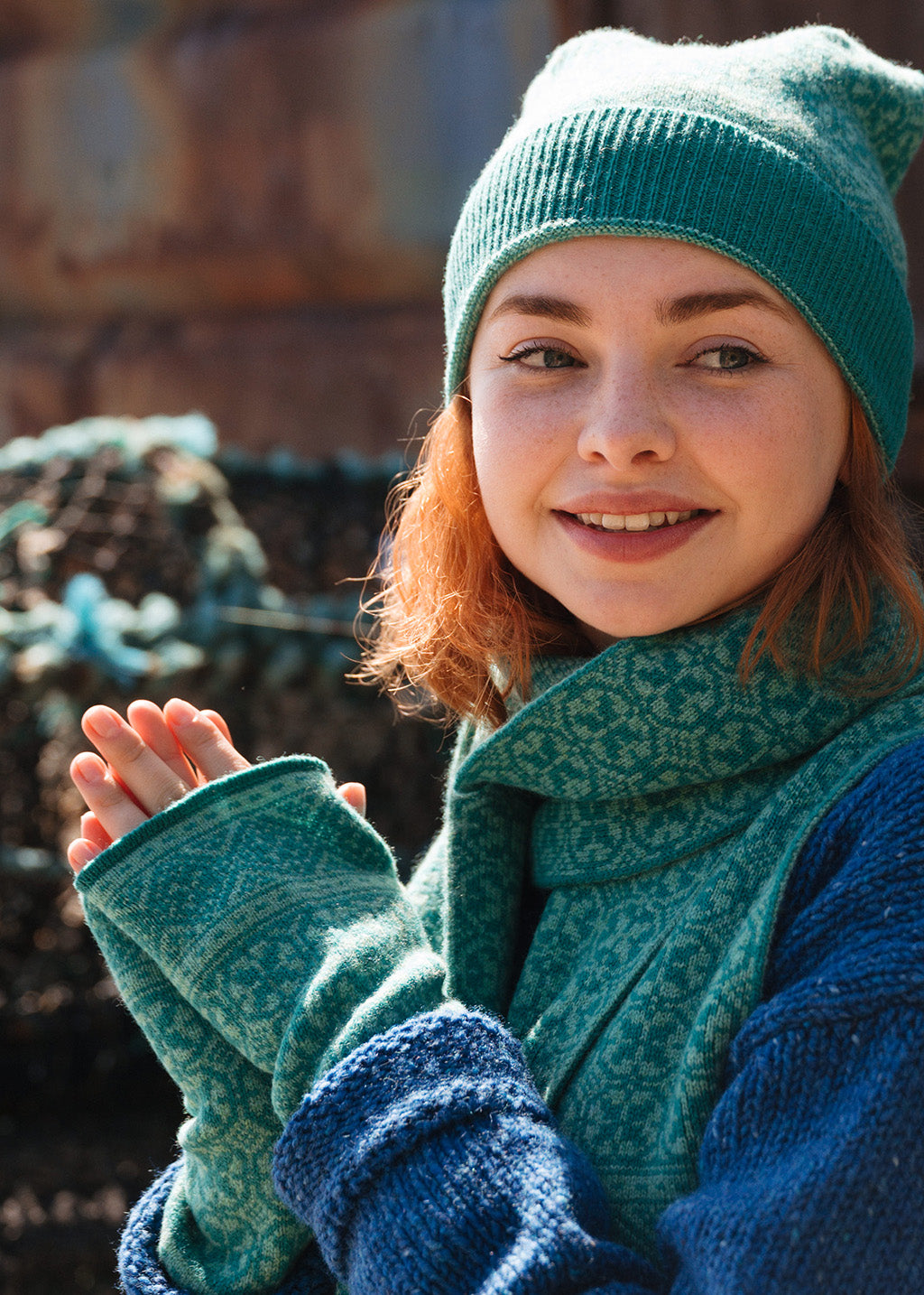 A smiling woman wearing a pair of sea green knit patterned wrist warmers, and matching knit scarf and pom pom hat.