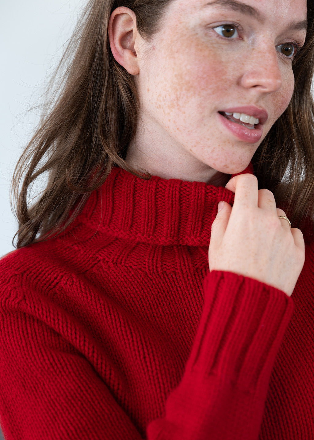 A close-up of a woman wearing a vibrant red wool knit polo neck jumper, touching the collar with her hand against a clean white background.