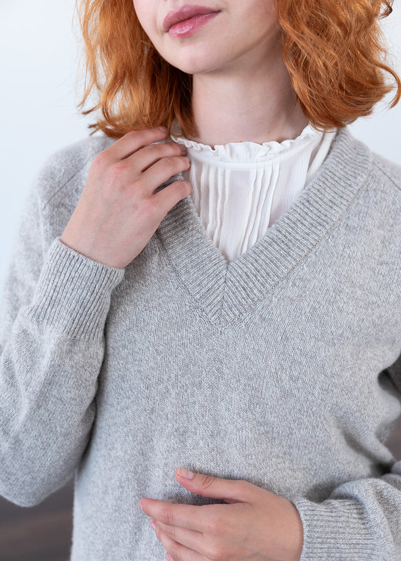A close-up of a woman wearing a light grey marl lambswool knit V-neck jumper over a white ruffled blouse. Her red hair is visible, and she is adjusting the collar of the sweater with one hand.