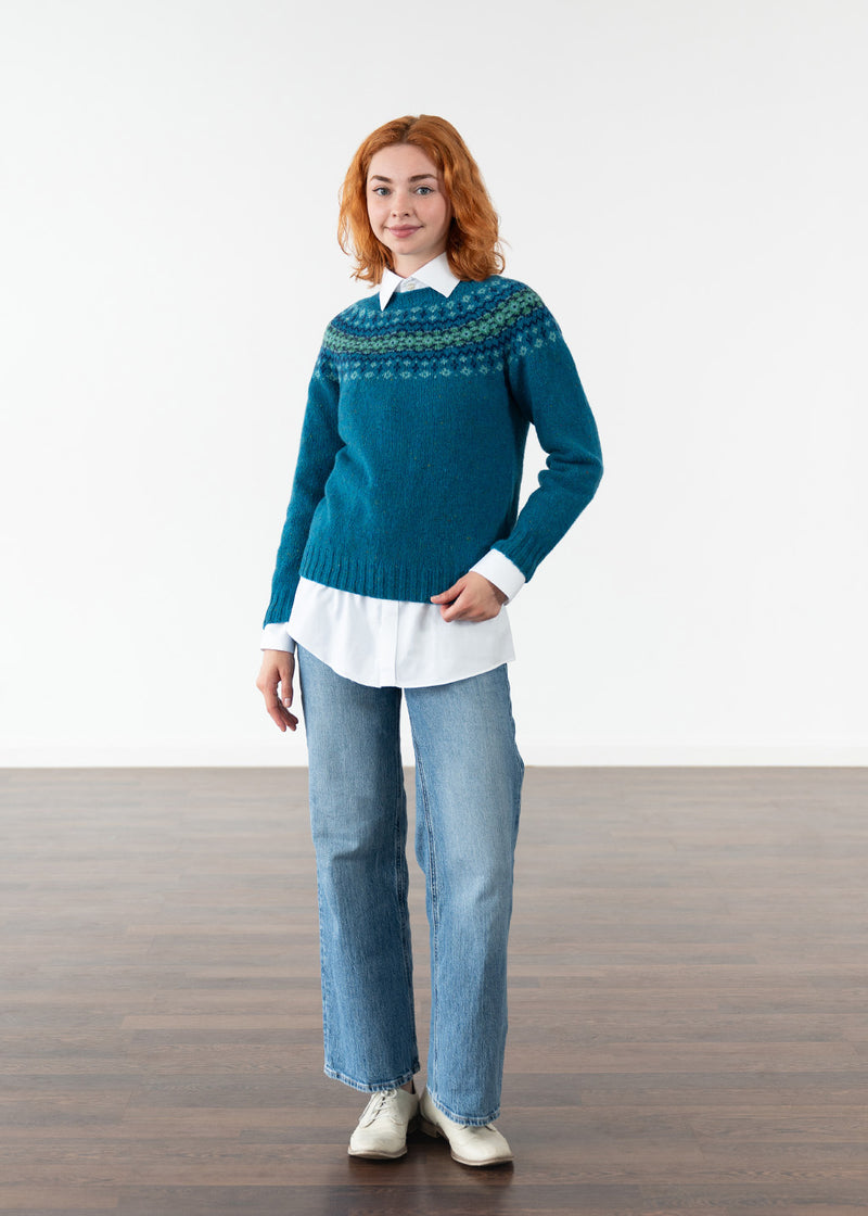 A woman modeling a teal blue Fair Isle knit jumper over a white collared shirt and light blue jeans, standing against a white studio wall on a dark wood floor.