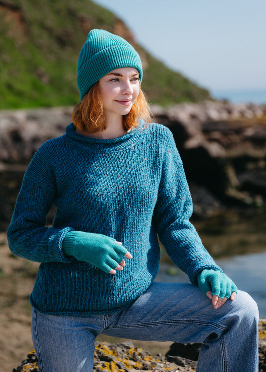 A woman wearing a teal blue chunky knit sweater, matching beanie, and fingerless gloves, standing by rocks by the sea with a grassy hill in the background.