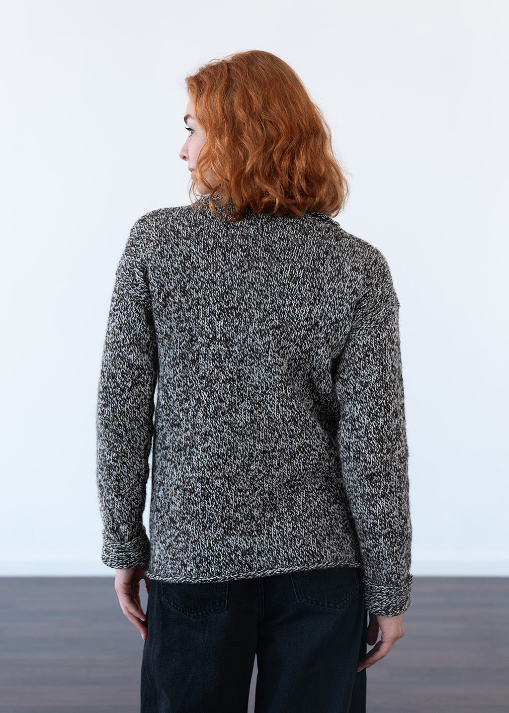 A woman with red hair is shown from the back wearing a black and white marled knit sweater and black jeans, standing against a plain white wall.