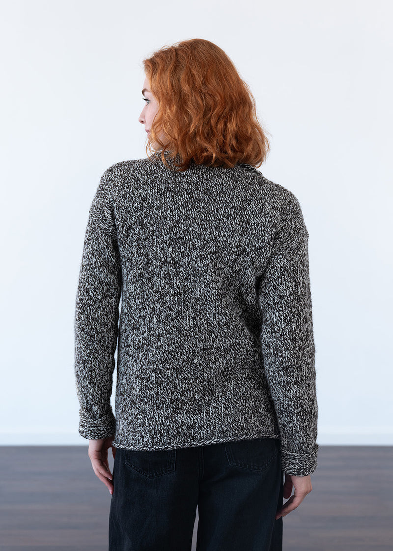 A woman with red hair is shown from the back wearing a black and white marled knit sweater and black jeans, standing against a plain white wall.