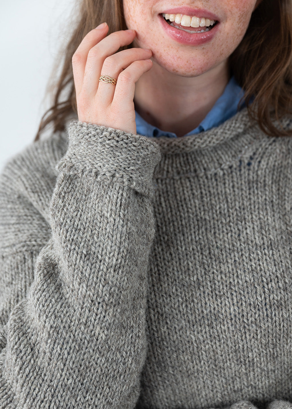 Close-up of a person wearing a chunky knit grey-brown undyed wool jumper over a light blue collared shirt, with one hand near the face and a gold ring on the finger.