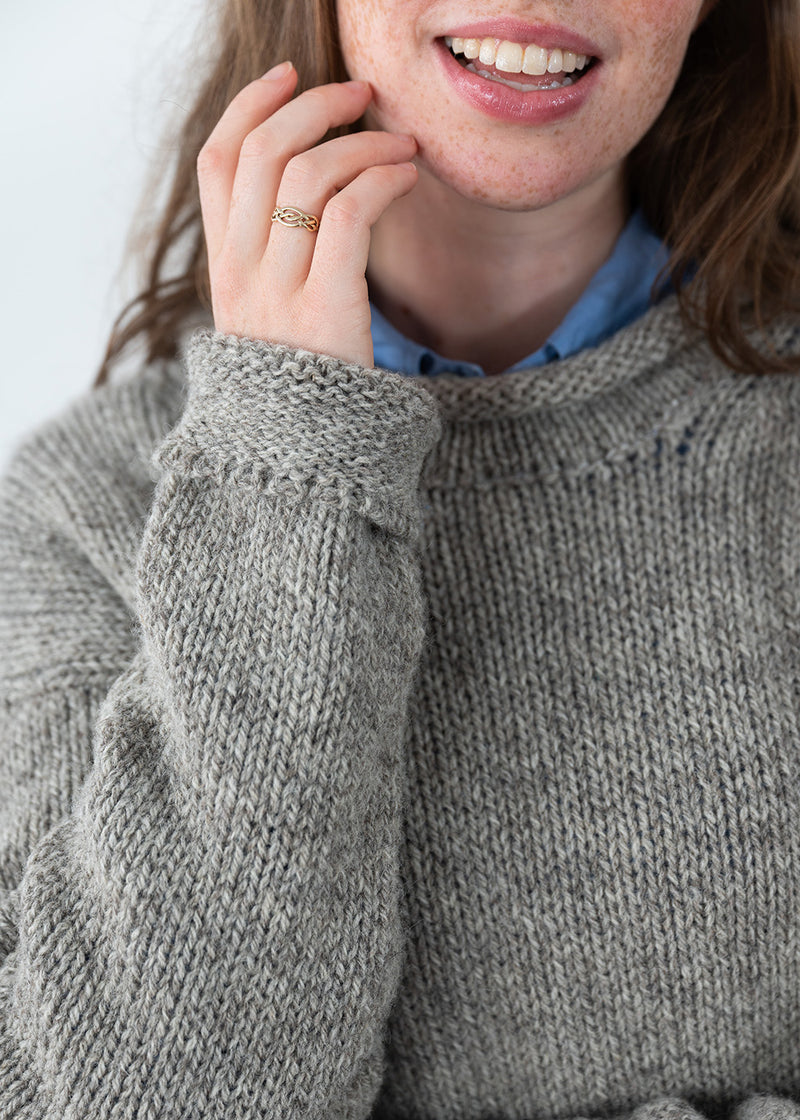 Close-up of a person wearing a chunky knit grey-brown undyed wool jumper over a light blue collared shirt, with one hand near the face and a gold ring on the finger.