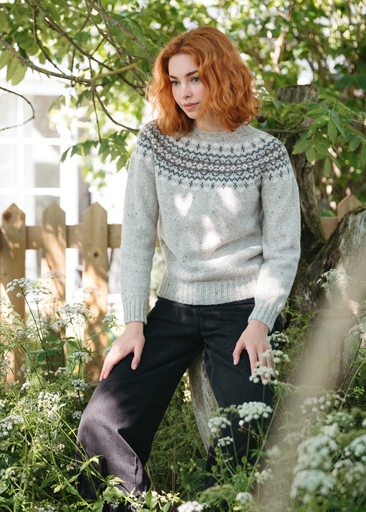 A woman with red hair wearing a light grey knit sweater with a dark grey Fair Isle yoke pattern and black jeans, posing outdoors in a garden setting with cow parsley foliage and a wooden fence.
