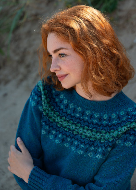 A woman with shoulder-length red hair wearing a teal blue and green fair isle knit sweater, photographed against a soft sandy background.