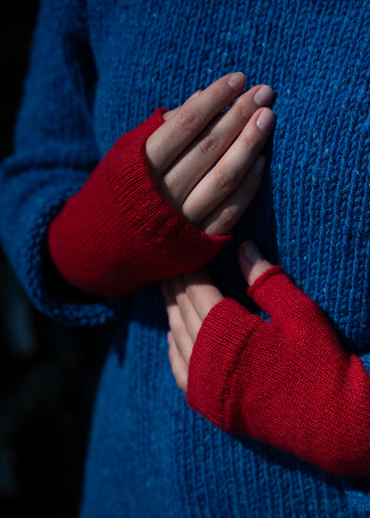 Red fingerless knit wrist warmer gloves worn on a person's hands, shown against a blue jumper in a close-up shot.