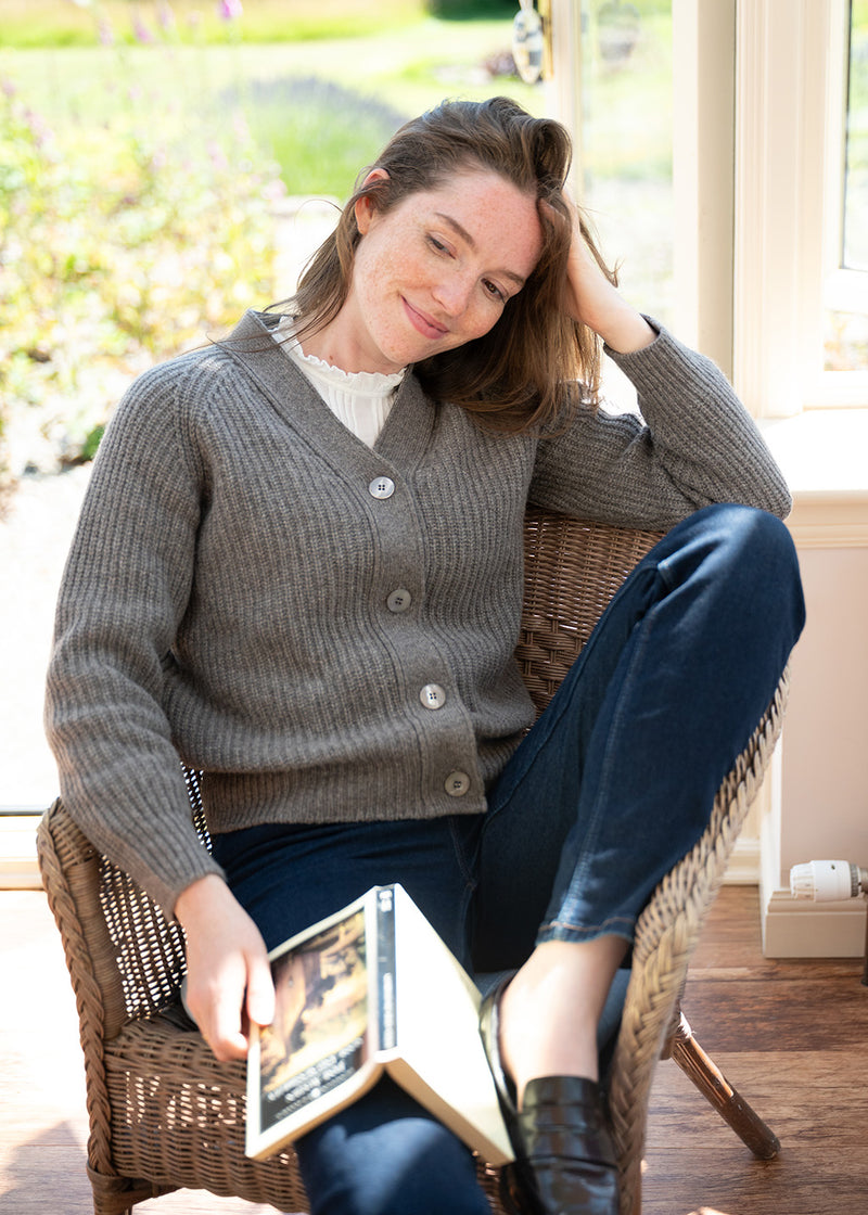 A woman sits in a wicker chair near an open glass door, wearing a brown ribbed button-front cardigan over a white top and dark blue jeans, holding a book.
