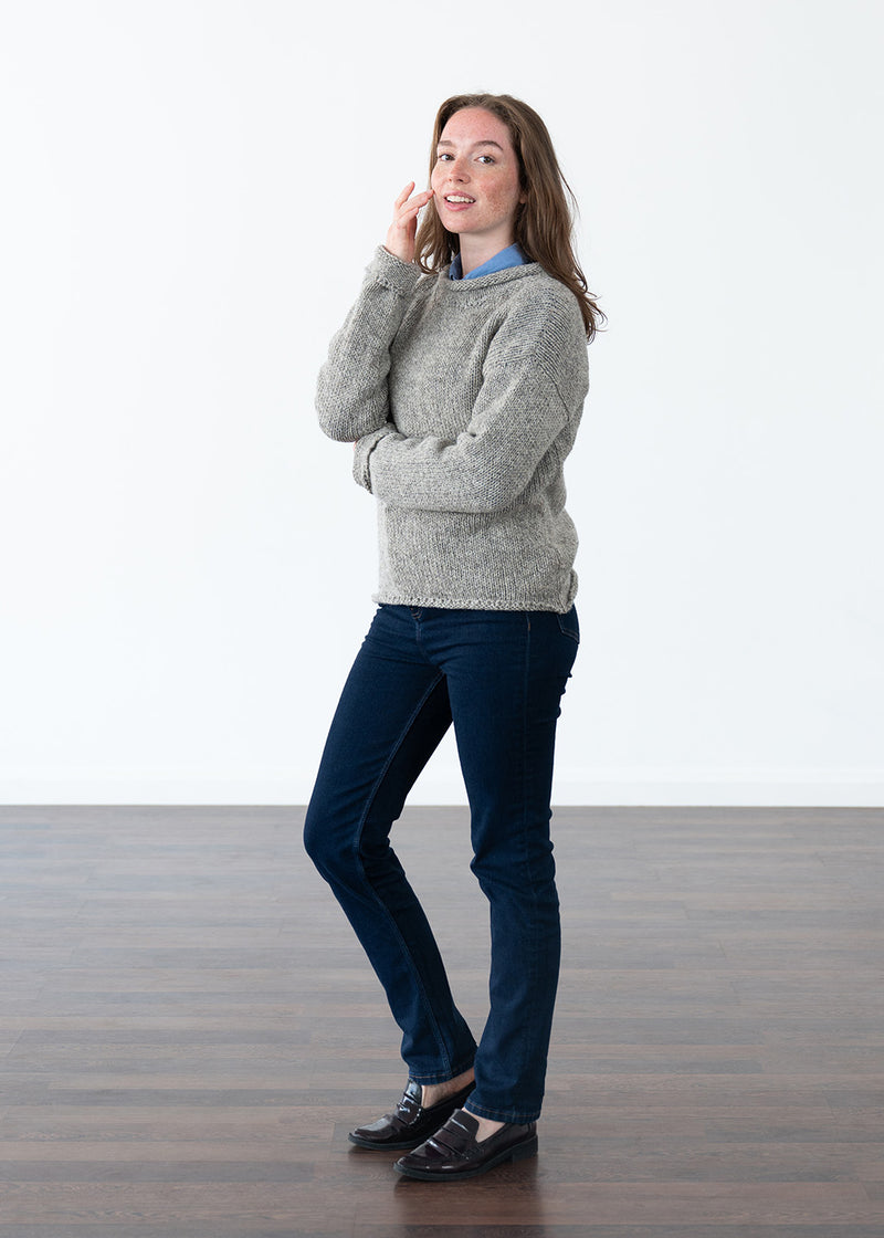 A woman stands in a minimalist studio with a white wall and dark wood floor, wearing a light grey knit jumper over a light blue collared shirt, dark navy jeans, and black loafers.