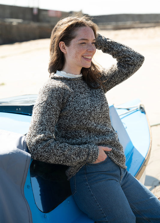 A woman wearing a marled ecru and black undyed wool knit jumper with blue jeans, sitting on a blue boat outdoors on a sunny day.