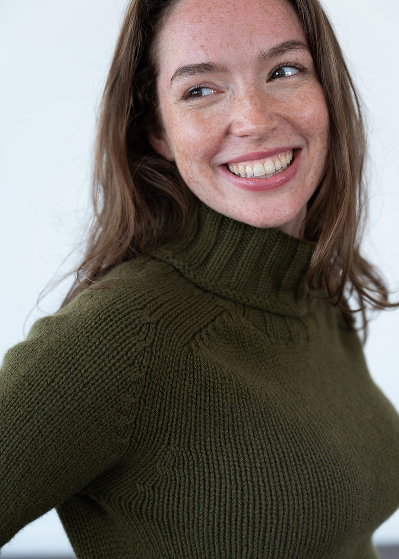 A smiling woman with brown hair wearing an olive green polo neck jumper, photographed against a plain light background.