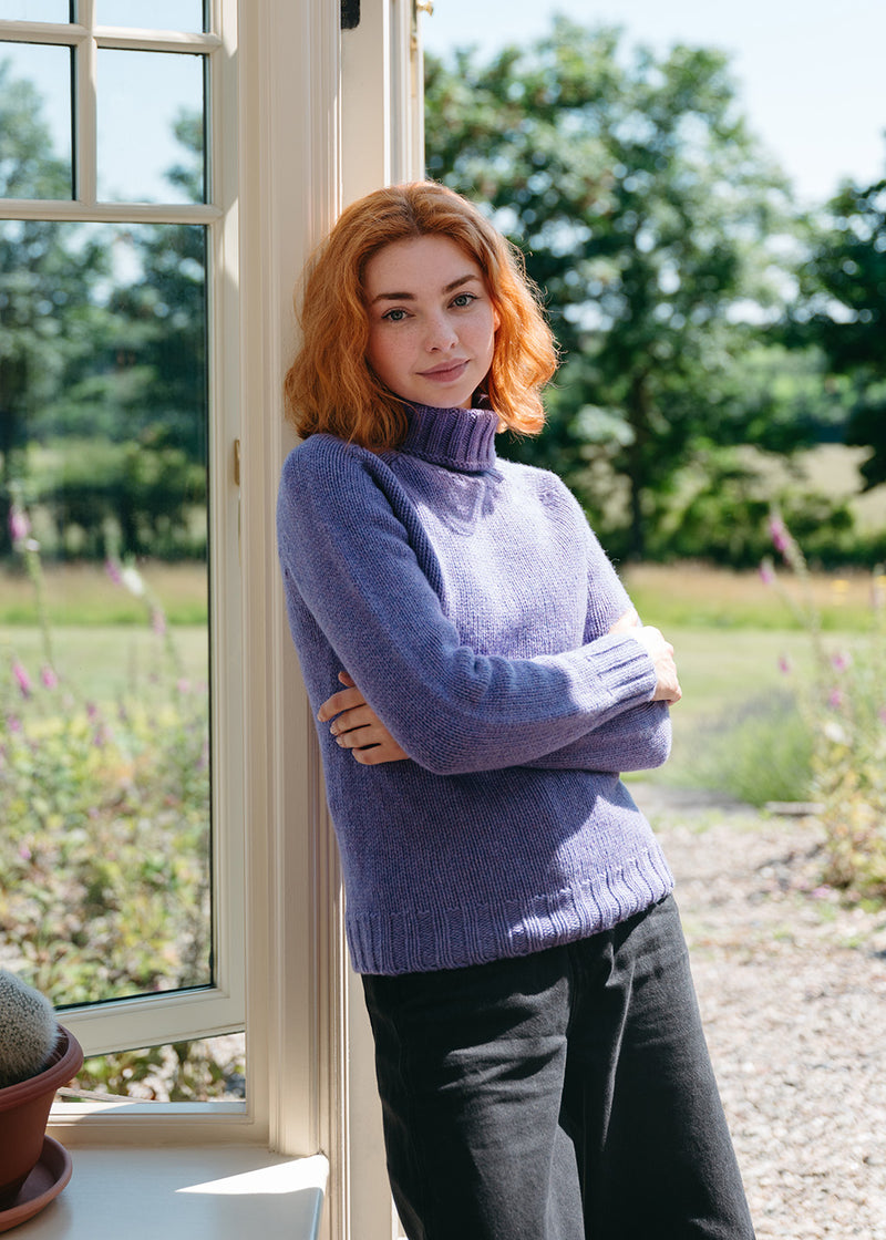 A woman with red hair stands leaning against a cream-framed window in a garden setting, wearing a lavender-purple ribbed polo neck jumper and black jeans.