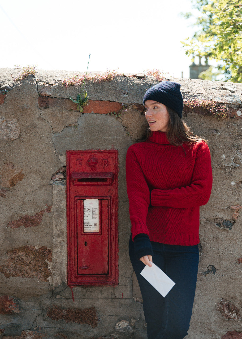 A woman in a red polo neck jumper, navy beanie, and dark jeans stands next to a red post box against a weathered stone wall, holding a white envelope.