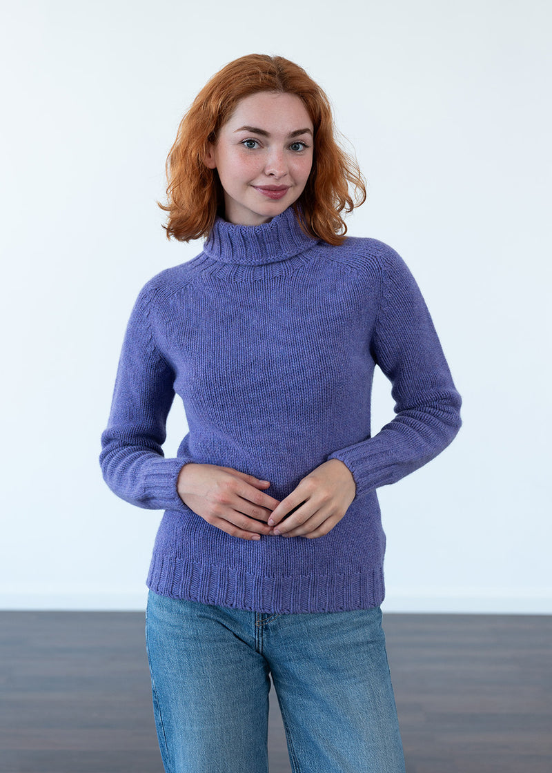 A woman with red hair wearing a purple polo neck jumper and blue jeans, standing against a plain white wall.
