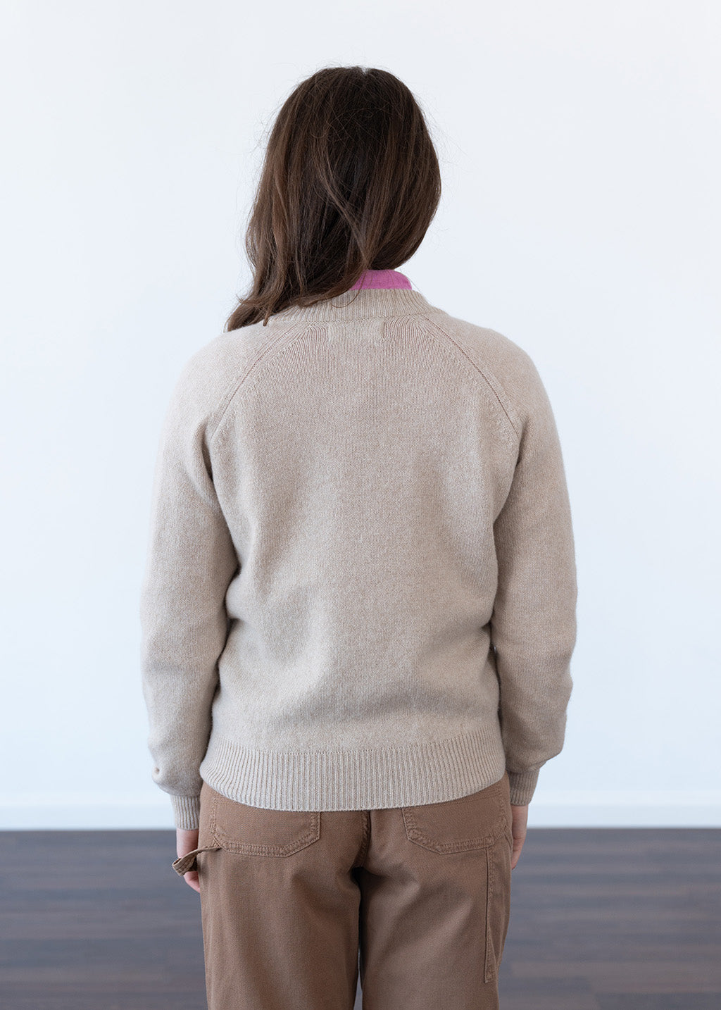 A woman viewed from behind wearing a light beige knit jumper and brown trousers, standing against a plain white wall.
