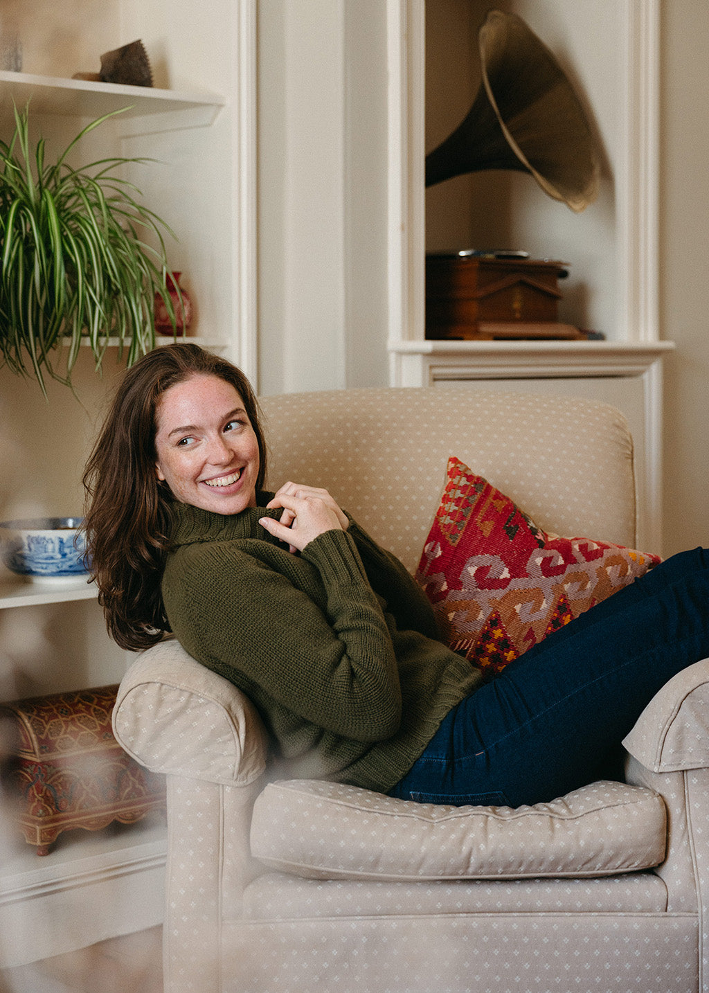 A woman smiling while reclining on a beige armchair in a cozy room, wearing an olive green knit polo neck jumper and dark blue jeans. A patterned red and orange throw cushion rests beside her.