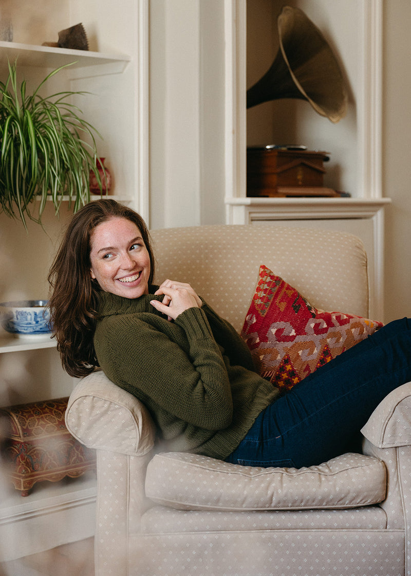 A woman smiling while reclining on a beige armchair in a cozy room, wearing an olive green knit polo neck jumper and dark blue jeans. A patterned red and orange throw cushion rests beside her.