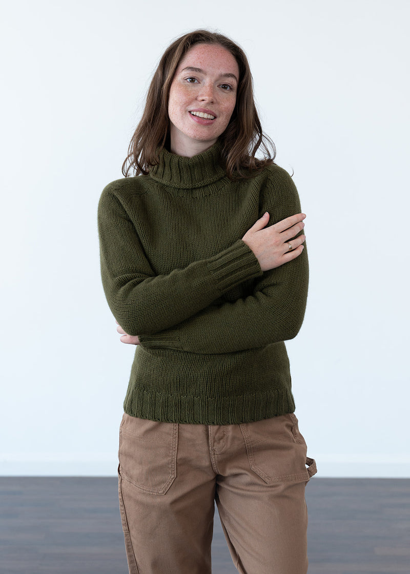 A woman wearing an olive green polo neck jumper and tan cargo trousers, standing with her arms crossed against a clean white studio background.