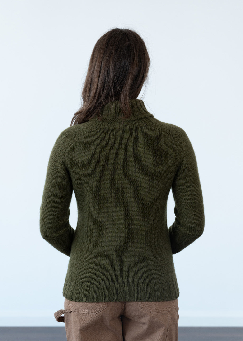 A woman viewed from behind wearing an olive green ribbed polo neck jumper and tan trousers against a plain white studio background.