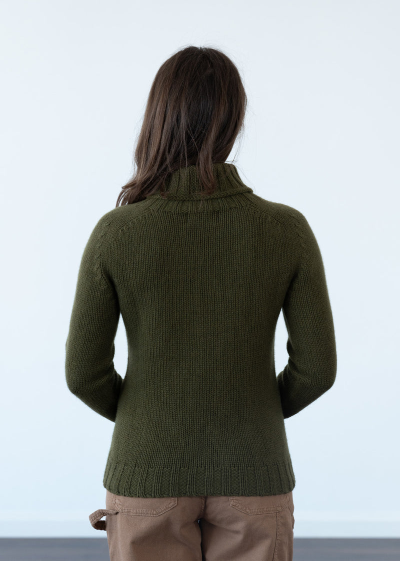A woman viewed from behind wearing an olive green ribbed polo neck jumper and tan trousers against a plain white studio background.