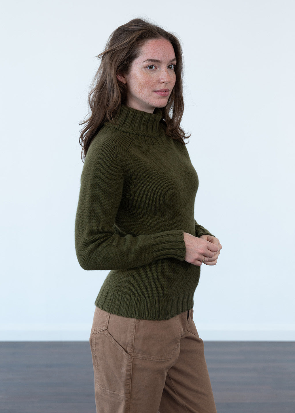 A woman wearing an olive green ribbed turtle neck sweater and tan cargo pants, standing against a white studio background.