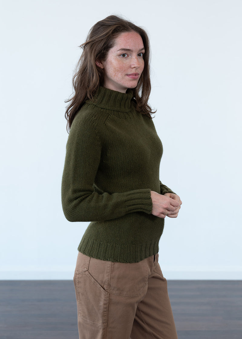 A woman wearing an olive green ribbed turtle neck sweater and tan cargo pants, standing against a white studio background.