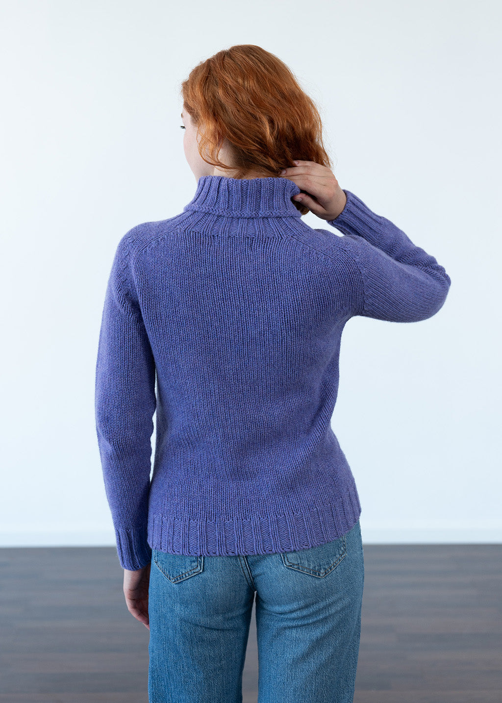 A woman with red hair is shown from behind wearing a ribbed purple turtleneck sweater and blue jeans, standing against a plain white wall.