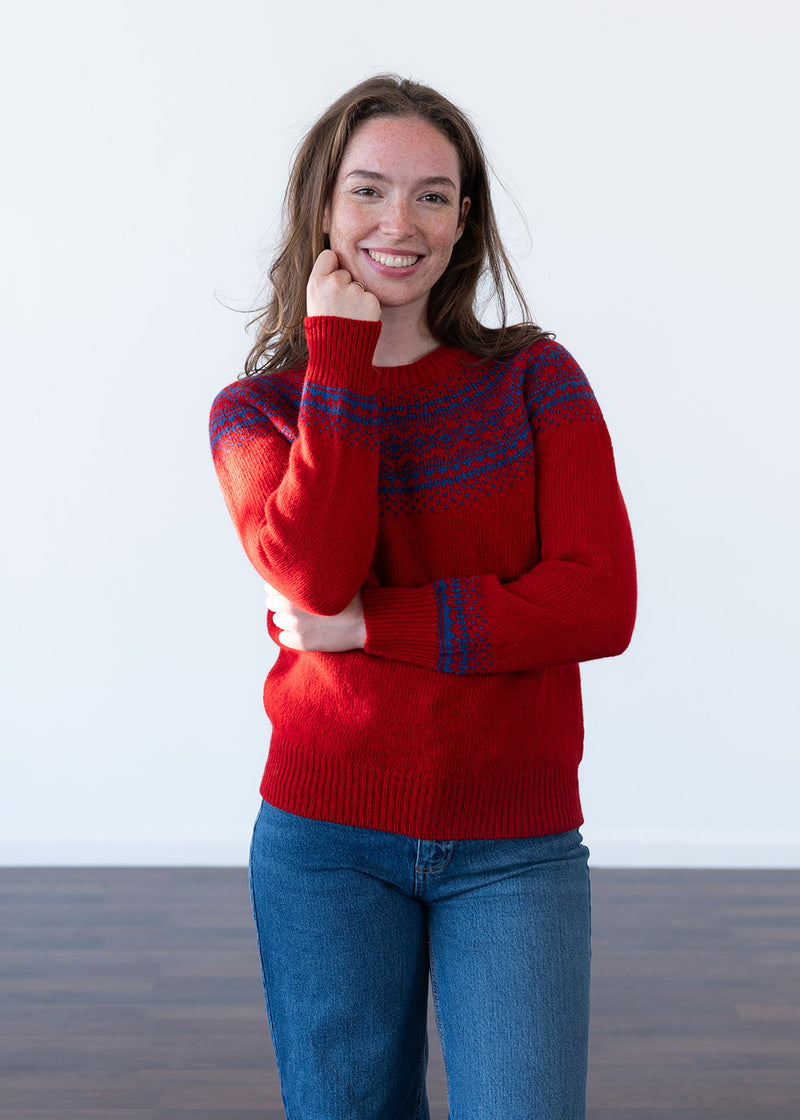 A smiling woman wearing a red knit sweater with blue patterned fair isle across the chest and cuffs, paired with blue jeans, standing against a plain white wall.