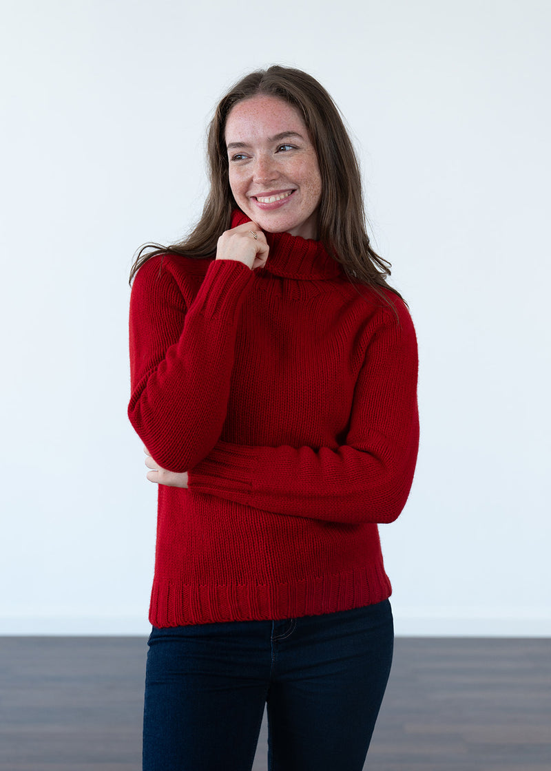 A smiling woman with long brown hair wearing a bright red ribbed polo neck jumper and dark blue jeans, standing against a plain white wall.