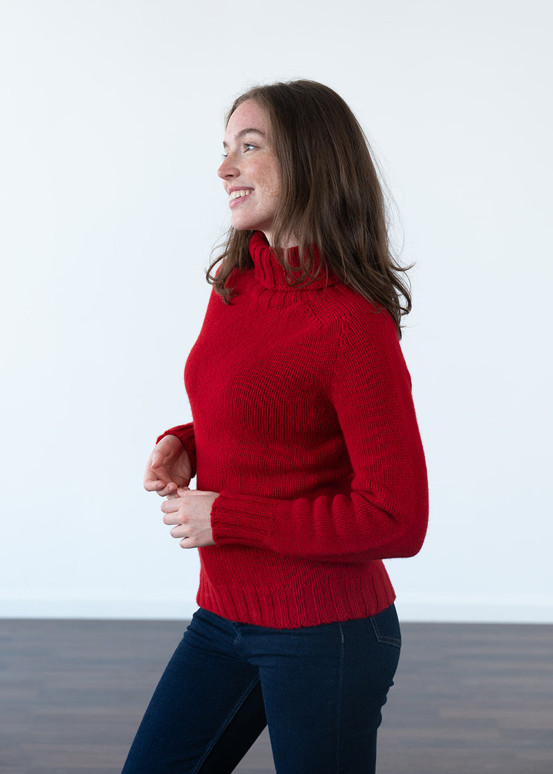 A woman wearing a bright red ribbed turtleneck sweater and dark blue jeans, standing against a clean white wall with a dark wooden floor.