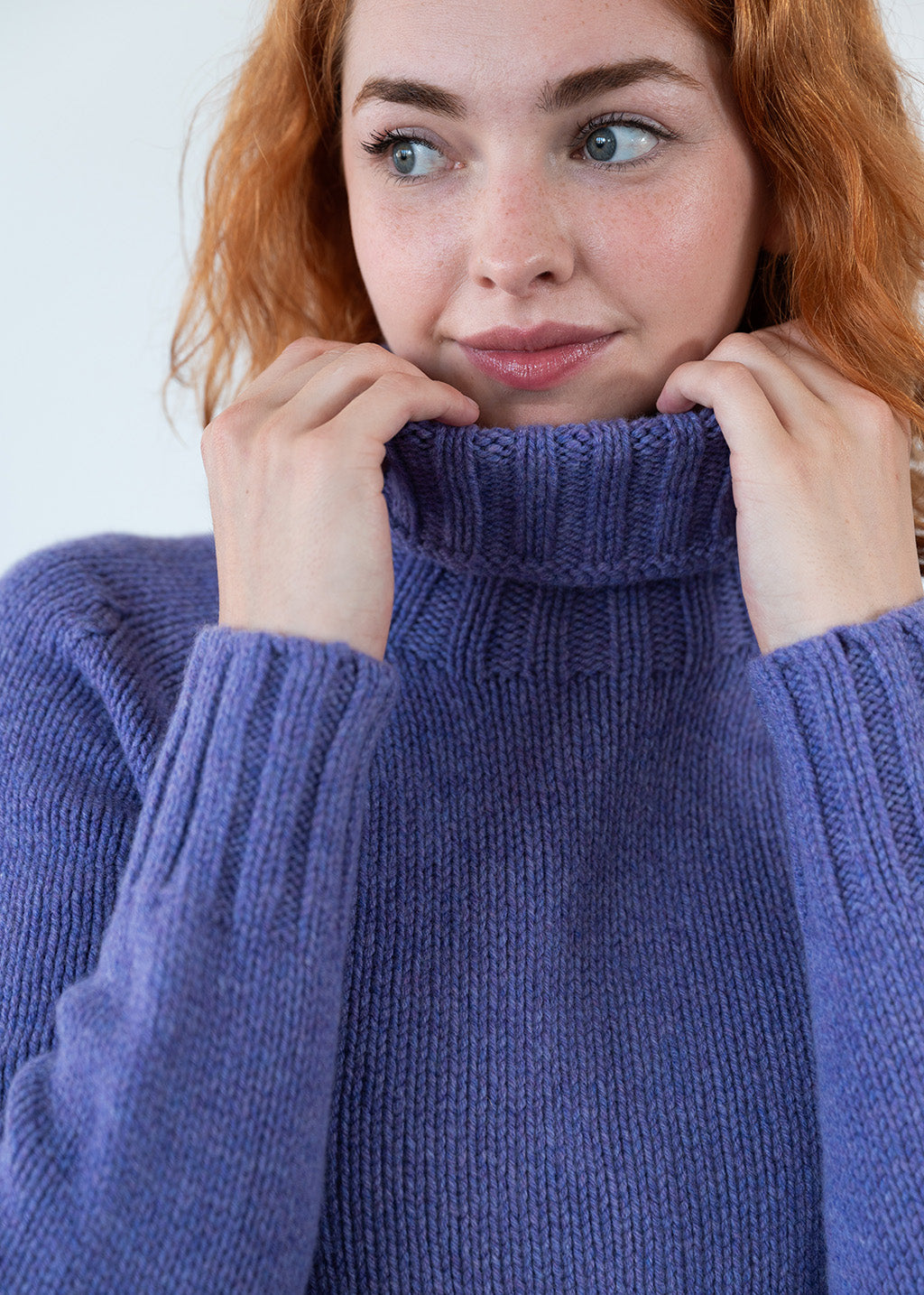 A close-up of a woman with red hair wearing a knit polo neck jumper in a heather purple colour, holding the collar up with both hands.