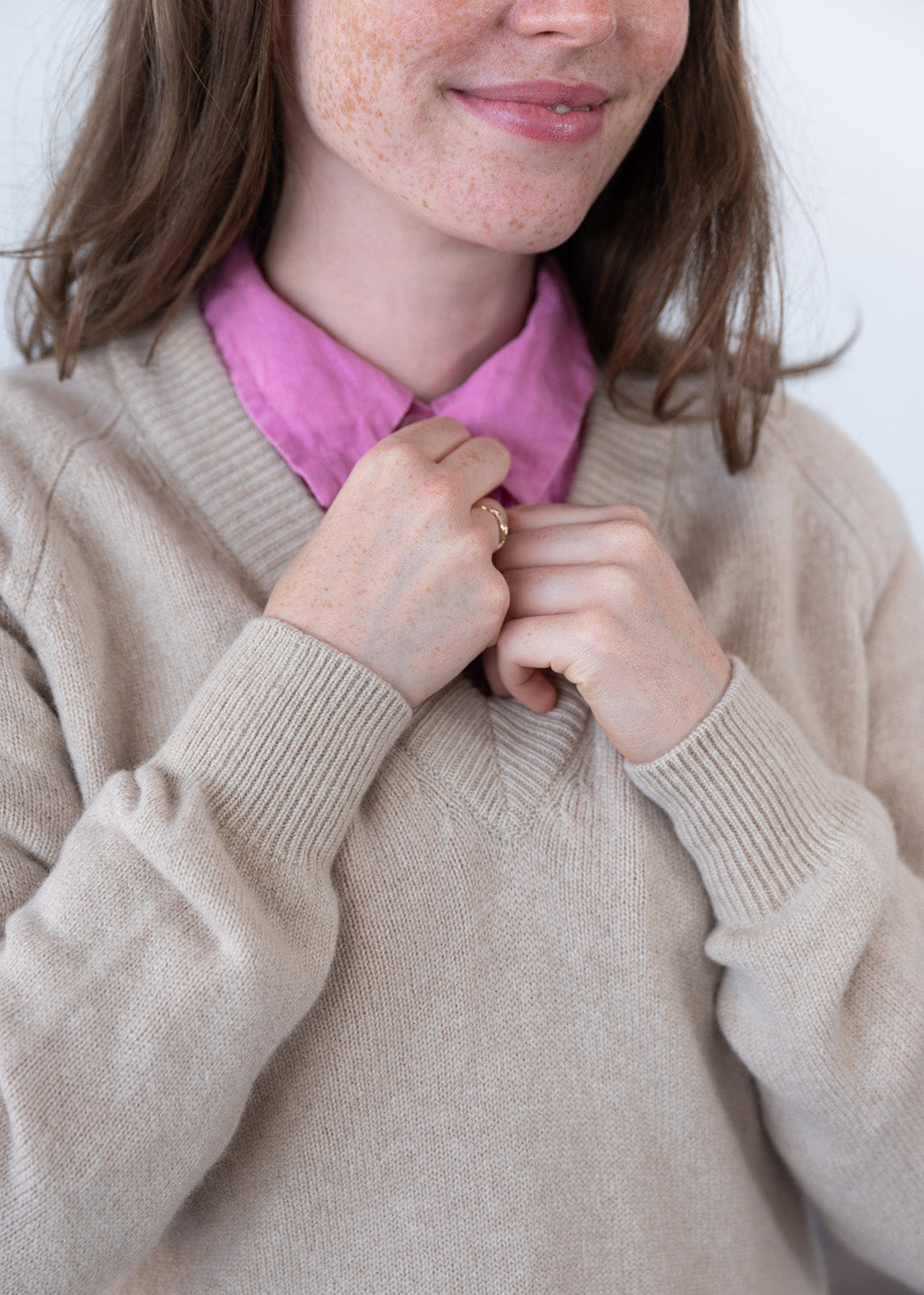 A close-up of a person wearing a beige knit lambswool sweater over a bright pink collared shirt, with hands adjusting the collar area.