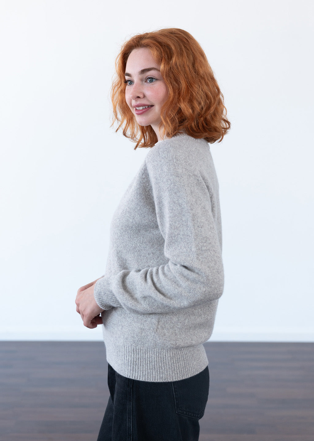 A woman with red hair wearing a light gray marl knit sweater and dark pants, standing in a minimalist studio setting with a white wall and dark wood floor.