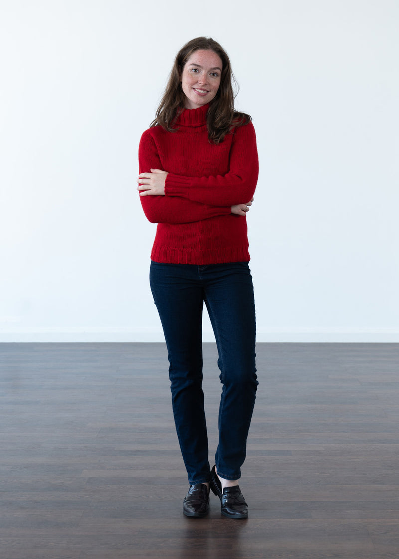 A woman stands with arms crossed wearing a bright red polo neck knit jumper, dark navy jeans, and black shoes, photographed in a minimalist studio with a white wall and dark wood floor.