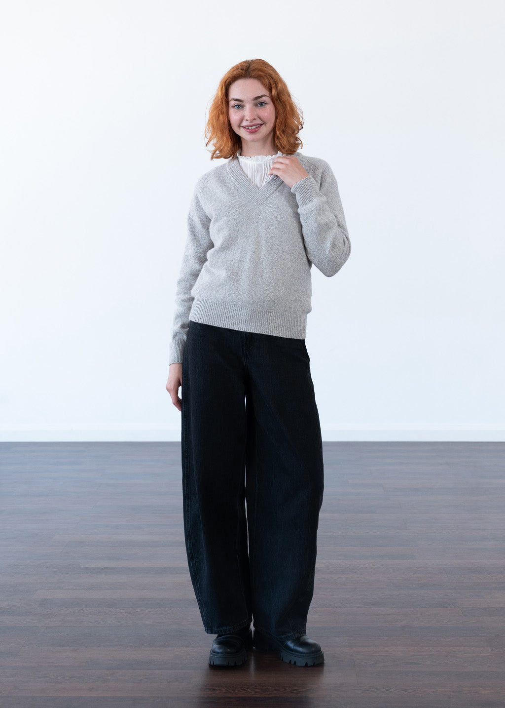 A woman with red hair stands in a minimalist studio setting with a white wall and dark wood floor, wearing a light grey marl knit V-neck sweater over a white top, paired with wide-leg black trousers and black shoes.