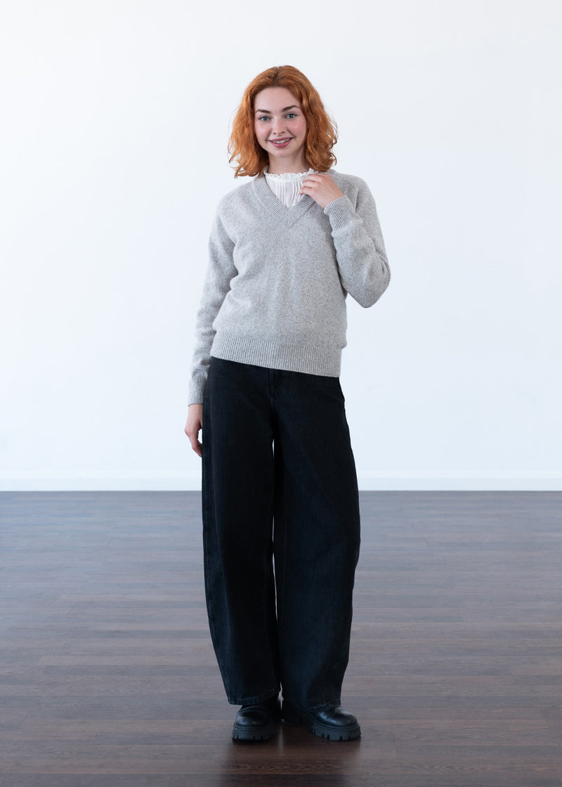 A woman with red hair stands in a minimalist studio setting with a white wall and dark wood floor, wearing a light grey marl knit V-neck sweater over a white top, paired with wide-leg black trousers and black shoes.