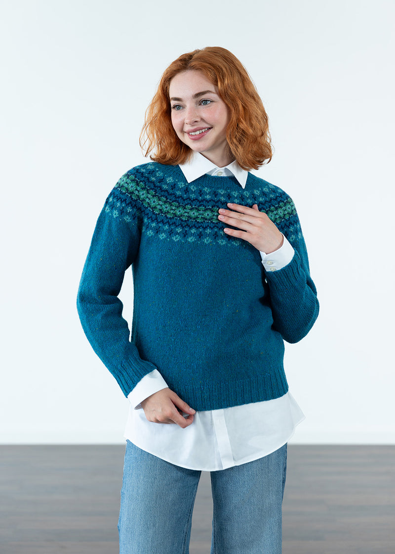 A woman with red hair wearing a teal blue Fair Isle knit jumper over a white collared shirt and blue jeans, standing against a clean white studio background.