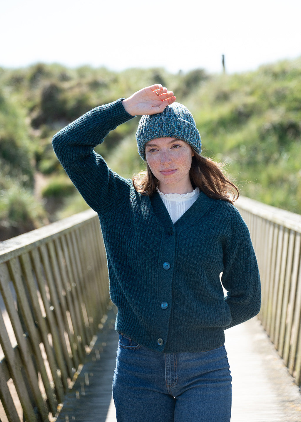 A woman walks along a wooden bridge with sand dunes behind, wearing a teal ribbed button-front cardigan over a white top with blue jeans and a marled teal beanie hat.