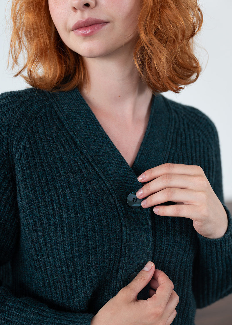 A close-up of a woman wearing a dark teal ribbed knit cardigan sweater with a button closure. The image shows her from the nose down, with her hands adjusting the front button of the sweater.