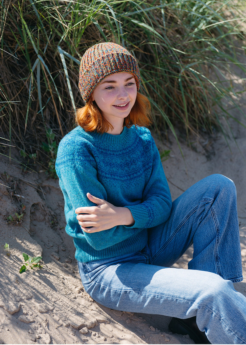 A woman sitting on sandy dunes wearing a teal fair isle sweater, blue jeans, and an orange marl beanie, with tall grass behind her.