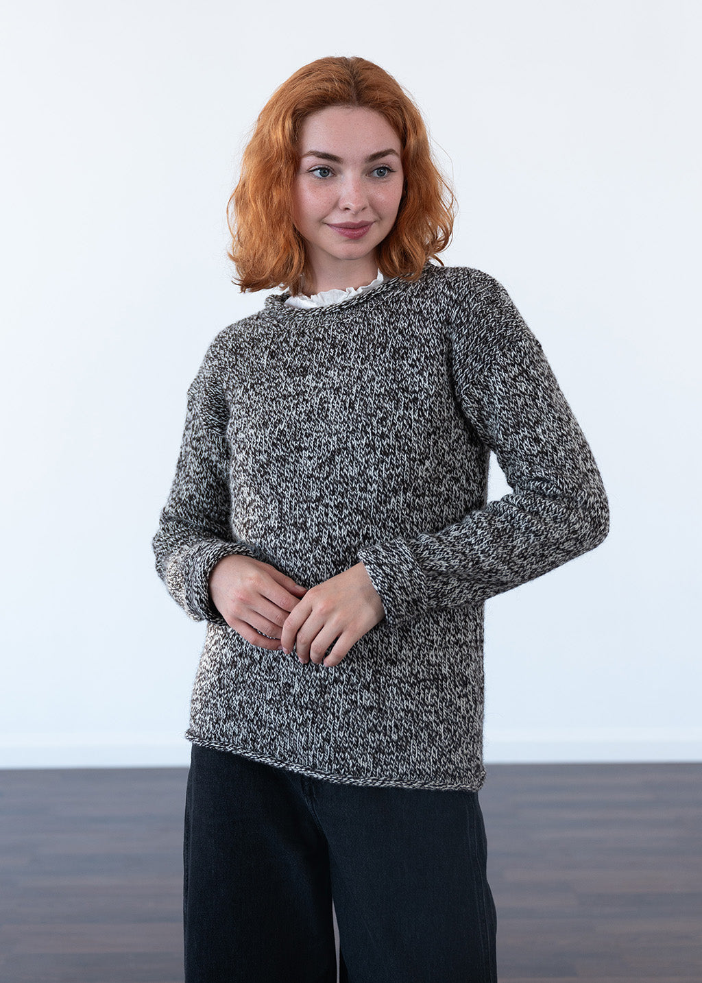 A woman with red hair wearing a textured black and white marled knit sweater and black jeans, standing against a plain white wall.