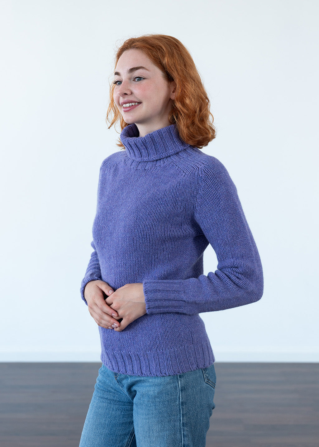 A woman with red hair wearing a lavender-purple turtleneck sweater and blue jeans, standing against a white wall.