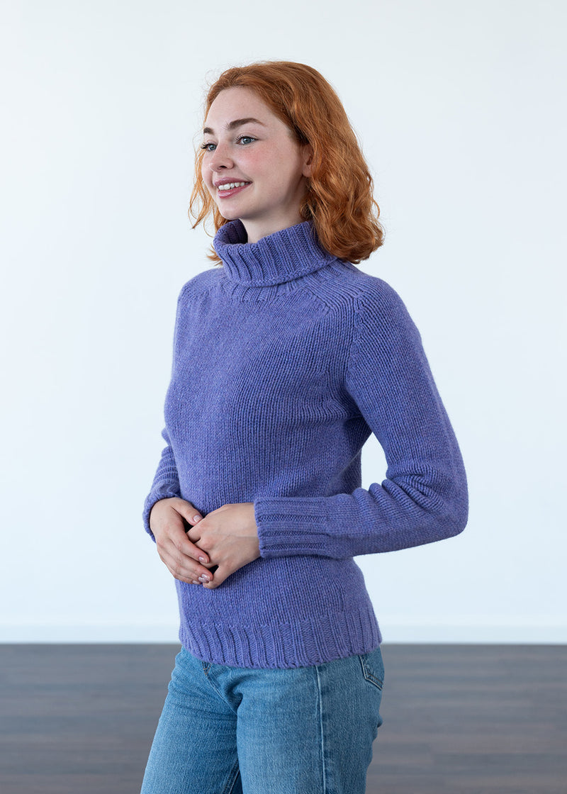 A woman with red hair wearing a lavender-purple turtleneck sweater and blue jeans, standing against a white wall.
